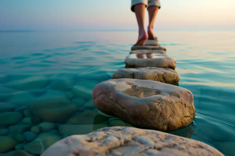 steps-to-sobriety-aa-recovery-living-sober-alcohol-detox-journey A close-up of a person walking on stepping stones over water, symbolizing the careful and deliberate steps taken in the journey to sobriety and recovery through AA.